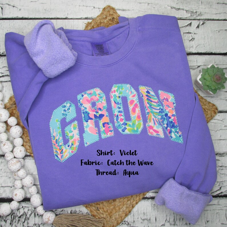 May include: A violet sweatshirt with the word "CROWN" appliqued across the front in a colorful floral pattern. The sweatshirt has a crew neck and long sleeves with a soft, fleece-lined interior. The text on the image says "Shirt: Violet, Fabric: Catch the Wave, Thread: Aqua."