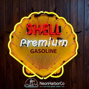Puede incluir: Un letrero de neón con forma de concha que presenta un fondo amarillo con el texto rojo SHELL, el texto blanco Premium y el texto rojo oscuro GASOLINE. El letrero está delineado con un tubo de neón amarillo brillante y herrajes negros.