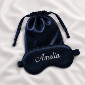 Könnte beinhalten: Eine dunkelmarineblaue Schlafmaske aus Satin mit dem in weißer Schreibschrift gestickten Namen Amelia, begleitet von einem passenden dunkelmarineblauen Satinbeutel mit Kordelzug, gerafftem Oberteil und zwei seitlichen Bändern.