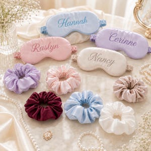 May include: A collection of satin sleep masks and scrunchies. The masks are light blue with 'Hannah', pink with 'Raslyn', purple with 'Corinne', and cream with 'Nancy' embroidered. The scrunchies are purple, pink, burgundy, light blue, cream, and tan.
