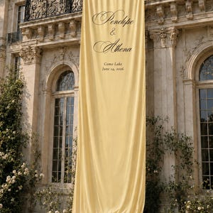 Può includere: Un lungo striscione in tessuto giallo pallido appeso a un balcone in pietra. Lo striscione presenta una scritta nera che recita Penelope & Athena, seguita dalla località Como Lake e dalla data 14 giugno 2026. Il tessuto ha un bordo arricciato.