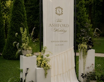 May include: A tall cream-colored fabric banner suspended on a black metal frame, featuring gold text reading The ASHFORD Wedding, SOFIA & EDWARD, and 22.06.2026. The banner has a ruffled diagonal hem and is flanked by white pedestals with floral arrangements.