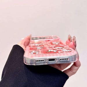 K&ouml;nnte beinhalten: Eine transparente Handyh&uuml;lle mit einem rosa-roten Karomuster und mehreren Hello Kitty-Illustrationen, darunter die Figur mit Erdbeeren. Die H&uuml;lle verf&uuml;gt &uuml;ber pr&auml;zise Aussparungen f&uuml;r den Ladeanschluss und die Lautsprecher an der Unterkante.
