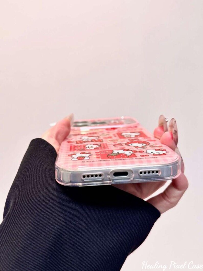 K&ouml;nnte beinhalten: Eine transparente Handyh&uuml;lle mit einem rosa-roten Karomuster und mehreren Hello Kitty-Illustrationen, darunter die Figur mit Erdbeeren. Die H&uuml;lle verf&uuml;gt &uuml;ber pr&auml;zise Aussparungen f&uuml;r den Ladeanschluss und die Lautsprecher an der Unterkante.