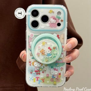 Puede incluir: Funda transparente para tel&eacute;fono con borde azul claro y agarre desplegable. La funda presenta un dise&ntilde;o de Hello Kitty con varias escenas y el texto "Kitty". El agarre desplegable tiene purpurina y peque&ntilde;os objetos flotantes.