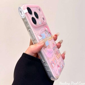 May include: A clear phone case with a pink camera lens protector and a repeating pattern of cartoon cat characters, carousel horses, and stars. The side of the case features small heart-shaped indentations along the edge and a white circular sticker.