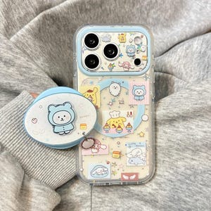 May include: A clear smartphone case with a light blue frame and a matching oval phone grip. The case features various cartoon character stickers, including a yellow dog in a chef hat, a white bear, and small star accents. The camera area is outlined in light blue.