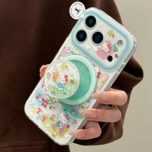 Puede incluir: Funda transparente para tel&eacute;fono con borde azul claro y pop socket. La funda presenta un dise&ntilde;o de Hello Kitty con un lazo rosa, mariposas y el texto "Kitty". El pop socket es verde menta y tiene una parte superior transparente con purpurina y un dise&ntilde;o de Hello Kitty.