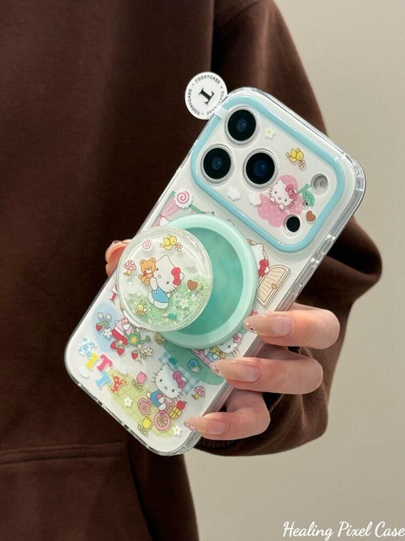 Puede incluir: Funda transparente para tel&eacute;fono con borde azul claro y pop socket. La funda presenta un dise&ntilde;o de Hello Kitty con un lazo rosa, mariposas y el texto "Kitty". El pop socket es verde menta y tiene una parte superior transparente con purpurina y un dise&ntilde;o de Hello Kitty.