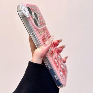 K&ouml;nnte beinhalten: Eine transparente Smartphone-H&uuml;lle mit rosa-wei&szlig;em Karomuster, kleinen Cartoon-Figuren und roten Herz-Symbolen. Die H&uuml;lle verf&uuml;gt &uuml;ber einen erh&ouml;hten rosa Kameraschutz und einen runden wei&szlig;en Aufkleber mit einem schwarzen Y-Symbol in der oberen Ecke.