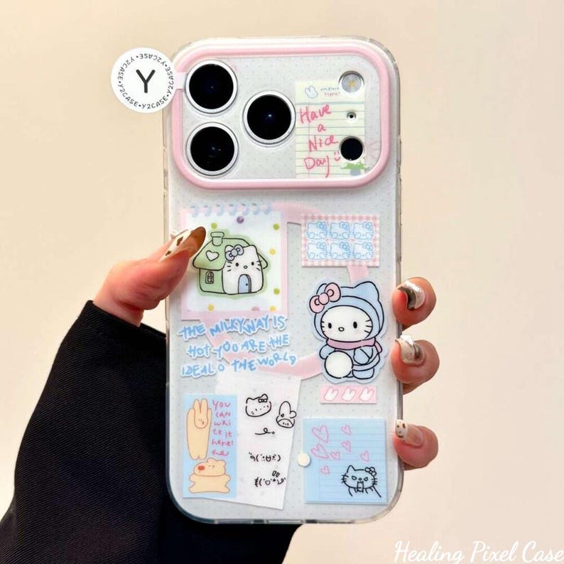 May include: A clear smartphone case featuring a pink camera frame and various stickers including a Hello Kitty character, a small house, a notepad with the text Have a Nice Day, and the phrase The Milkyway is hot you are the ideal of the world.