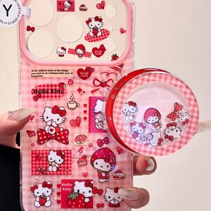 K&ouml;nnte beinhalten: Eine transparente Handyh&uuml;lle mit rosa Vichy-Karo-Hintergrund, verziert mit Hello Kitty-Grafiken, Erdbeeren und Herzen. Die H&uuml;lle enth&auml;lt einen runden Pop-Out-Griff mit schwimmenden Hello Kitty-Anh&auml;ngern sowie den Text, To be without some of the things you want is an indispensable part of happiness und In all the shabby fading, please shine forever.