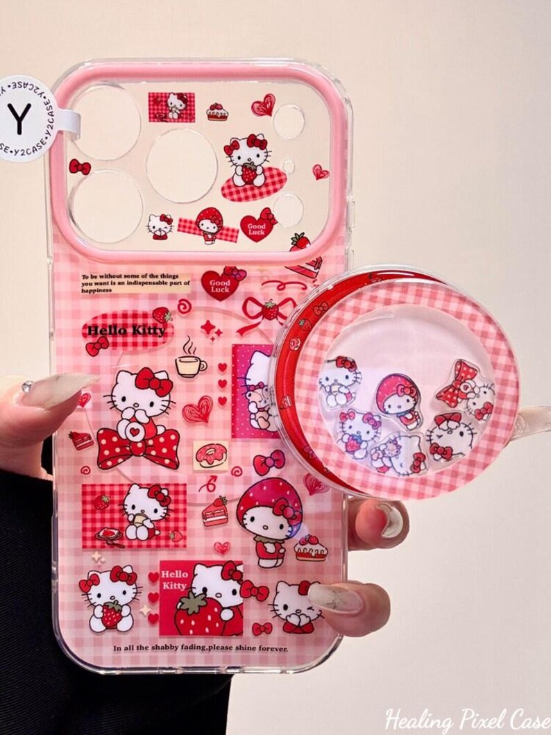 K&ouml;nnte beinhalten: Eine transparente Handyh&uuml;lle mit rosa Vichy-Karo-Hintergrund, verziert mit Hello Kitty-Grafiken, Erdbeeren und Herzen. Die H&uuml;lle enth&auml;lt einen runden Pop-Out-Griff mit schwimmenden Hello Kitty-Anh&auml;ngern sowie den Text, To be without some of the things you want is an indispensable part of happiness und In all the shabby fading, please shine forever.