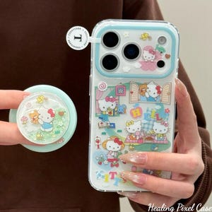 Puede incluir: Funda transparente para tel&eacute;fono con borde azul claro y un pop socket a juego. La funda presenta un colorido dise&ntilde;o de Hello Kitty con varias escenas. El pop socket tiene un interior brillante con un dise&ntilde;o de Hello Kitty y oso.