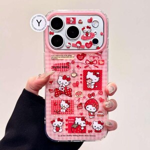 K&ouml;nnte beinhalten: Eine transparente Handyh&uuml;lle mit rosafarbenem Karomuster und zahlreichen Hello Kitty-Grafiken in Rot und Wei&szlig;. Das Design umfasst Erdbeeren, Herzen und kleine Textelemente. Ein rundes wei&szlig;es Etikett mit einem schwarzen Y-Logo ist oben befestigt.