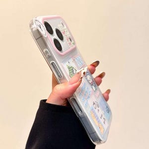 May include: A clear smartphone case with a light pink camera frame and a collage of various cartoon character stickers and blue text accents. The case features a transparent back panel that displays the phone's design underneath the decorative stickers.