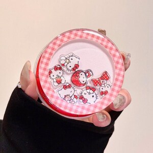 K&ouml;nnte beinhalten: Ein rundes, transparentes Kunststoffgeh&auml;use mit einem rosa-wei&szlig; karierten Rand. Im Inneren befinden sich mehrere kleine, lose Acryl-Anh&auml;nger, die Hello Kitty-Figuren in verschiedenen Posen darstellen, darunter eine mit einer Erdbeere und eine mit einer roten Schleife.