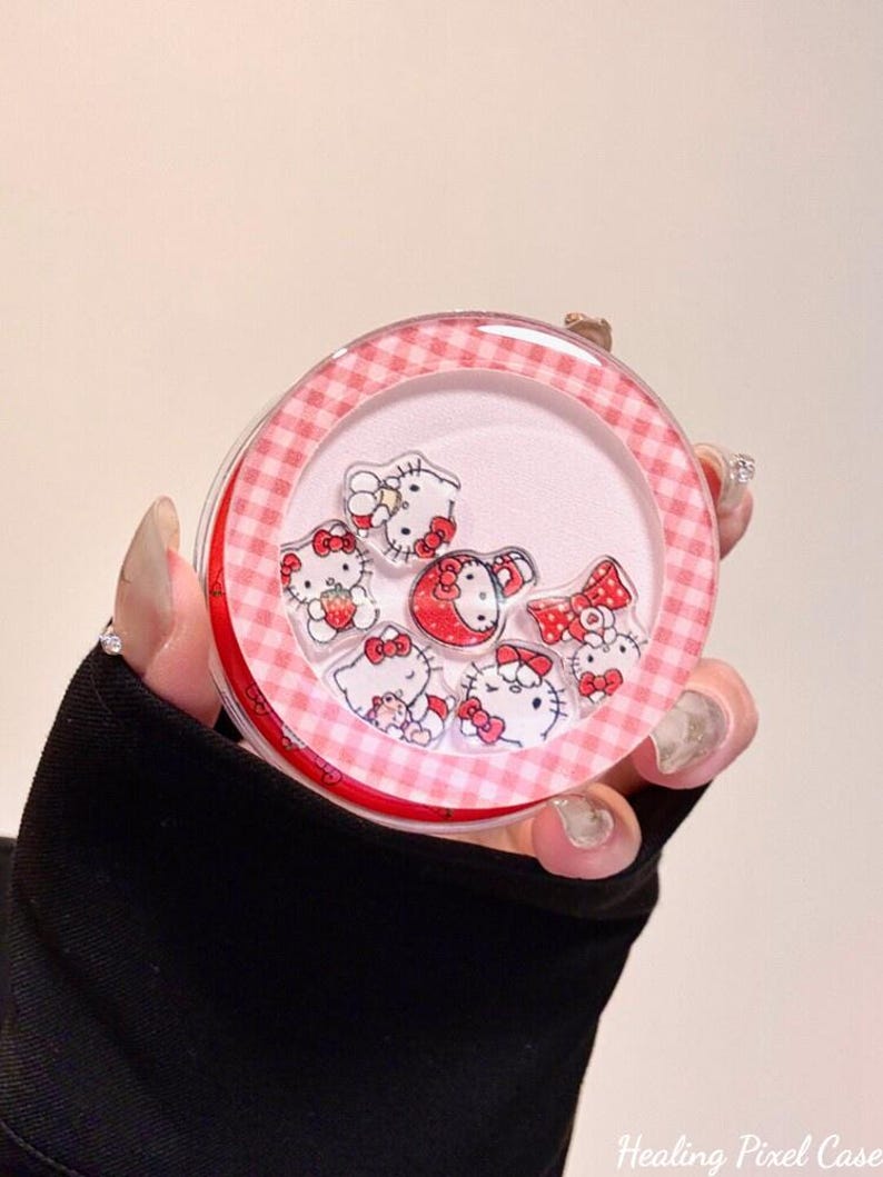K&ouml;nnte beinhalten: Ein rundes, transparentes Kunststoffgeh&auml;use mit einem rosa-wei&szlig; karierten Rand. Im Inneren befinden sich mehrere kleine, lose Acryl-Anh&auml;nger, die Hello Kitty-Figuren in verschiedenen Posen darstellen, darunter eine mit einer Erdbeere und eine mit einer roten Schleife.