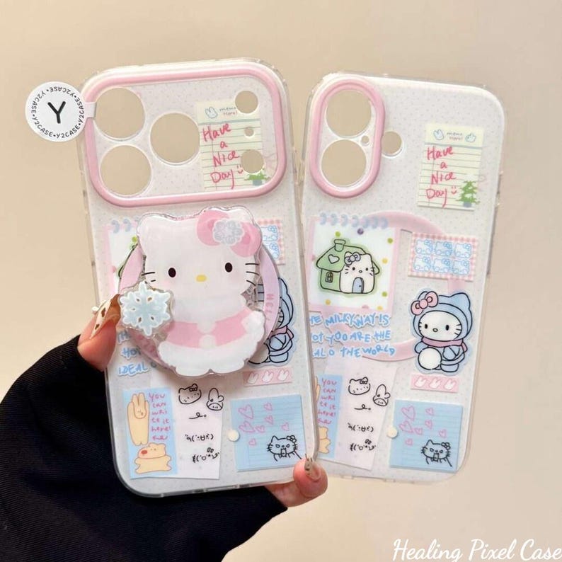 May include: Two clear phone cases with pink camera borders, decorated with various stickers including Hello Kitty, small houses, and notes that say Have a Nice Day. One case features a circular Hello Kitty stand with a snowflake accent.