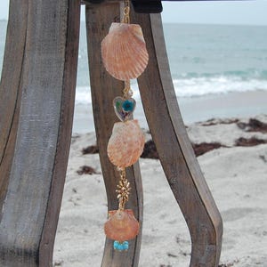 May include: A hanging car charm featuring three light orange scallop shells, a small heart-shaped pendant containing a blue flower, a gold-toned sun-shaped metal charm, and a cluster of small turquoise beads, all connected by a gold-toned metal chain.