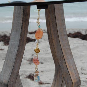 May include: A gold-toned metal chain charm featuring a collection of hanging elements including a large orange scallop shell, a white pearl, a small gold-toned metal shell, a pink textured shell, and a miniature glass bottle with a cork stopper.
