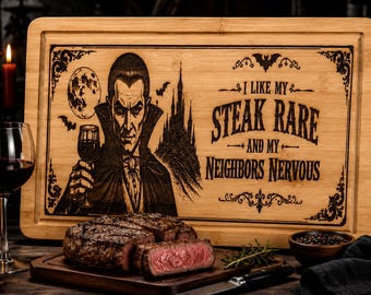 May include: A rectangular bamboo cutting board featuring a laser-engraved illustration of a vampire holding a wine glass, a castle, and the text: I LIKE MY STEAK RARE AND MY NEIGHBORS NERVOUS. The board has a grooved border and decorative corners.