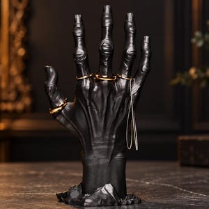 May include: A matte black hand-shaped jewellery display stand featuring elongated, skeletal-like fingers. The stand is adorned with four simple gold-toned rings on the fingers and a thin, delicate silver-toned chain draped from the ring finger.