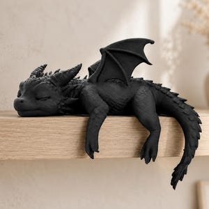 May include: A matte black dragon figurine in a sleeping position, featuring textured scales, folded wings, and a long tail that hangs over the edge. The creature has closed eyes, small horns on its head, and a smooth, detailed surface finish.