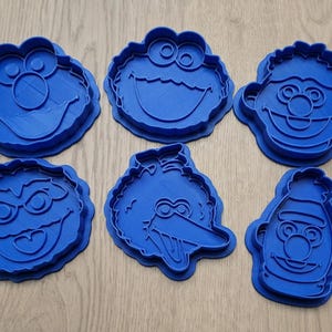May include: A set of six blue plastic cookie cutters featuring the faces of various Sesame Street characters, including Elmo, Cookie Monster, Bert, Ernie, Oscar the Grouch, and Big Bird, each with distinct facial features and expressions.