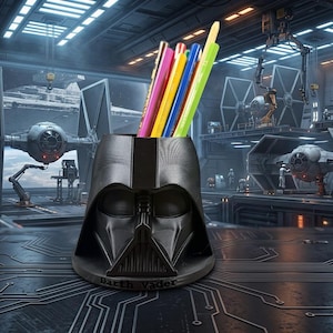 May include: A black pencil holder shaped like the helmet of Darth Vader, featuring a matte finish and the name Darth Vader embossed on the base. The holder contains several coloured pencils and is positioned in a dark, futuristic hangar setting.