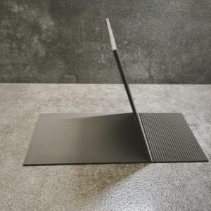 Op de afbeelding: Een zwarte metalen standaard met een gladde, platte rechthoekige basis verbonden met een verticaal, schuin steunpaneel. De basis bevat een getextureerd, geribbeld gedeelte aan de rechterkant, terwijl het verticale paneel een dunne, lichte rand heeft.