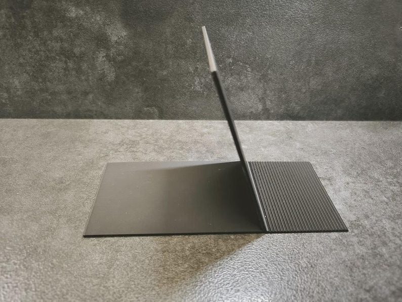 Op de afbeelding: Een zwarte metalen standaard met een gladde, platte rechthoekige basis verbonden met een verticaal, schuin steunpaneel. De basis bevat een getextureerd, geribbeld gedeelte aan de rechterkant, terwijl het verticale paneel een dunne, lichte rand heeft.