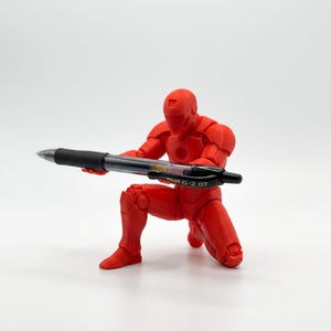 May include: A bright red, articulated humanoid figure in a kneeling pose, holding a black Pilot G-2 07 gel pen horizontally in its hands. The figure features a smooth, matte finish and a helmeted head, serving as a functional desk accessory.