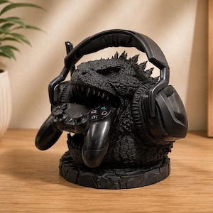 May include: A black monster-shaped stand featuring a textured, scaly surface and sharp teeth. The figure holds a black gaming controller in its mouth and supports a pair of black over-ear headphones on its head, all resting on a circular base.