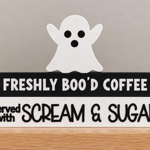 May include: A decorative stack of two rectangular blocks topped with a white ghost figure. The black block features the white text FRESHLY BOO'D COFFEE. The white block features the black text served with SCREAM & SUGAR in a mix of fonts.