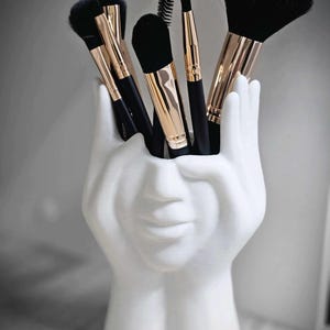 May include: A white, face-shaped makeup brush holder filled with various black and gold makeup brushes. The holder is a sculpted design of a face and hands, creating a unique and artistic storage solution for beauty tools.