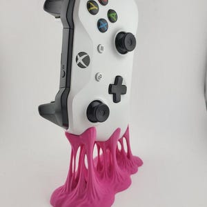 May include: A white Xbox controller with black detailing, displayed on a bright pink, artistic stand. The controller has coloured buttons and the Xbox symbol. The stand has a distinctive, dripping design, adding a playful aesthetic to the gaming accessory.