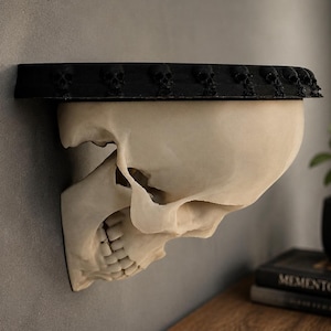 May include: A wall-mounted shelf featuring a beige human skull base and a flat black top surface. The edge of the black shelf is decorated with a repeating row of small, sculpted skulls. The structure is mounted against a plain grey wall.