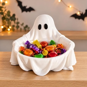 May include: A white ceramic bowl shaped like a ghost with two black oval eyes, filled with assorted sweets wrapped in colourful metallic foil. The ghost figure has draped fabric-like folds and serves as a container for the small treats.