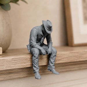May include: A monochromatic grey figurine of a cowboy wearing a wide-brimmed hat, a collared shirt, and trousers, depicted in a seated position with legs dangling over the edge. The figure has a holster at the hip and is posed in a slumped posture.