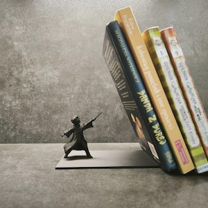 Op de afbeelding: Een donker metalen boekensteun met een klein figuurtje in een gewaad dat een toverstaf vasthoudt, ondersteunt vier boeken met de titels Panda z durej, Kubusiowy pamiętnik i inne opowieści en twee delen van Dziennik Cwaniaczka door Jeff Kinney.