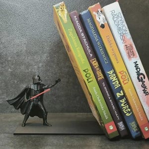 May include: A black metal bookend featuring a Darth Vader figure holding a red lightsaber, supporting a row of five books with colourful spines including titles like Zagubiona fotografia, Ekipa na tropie, Banda z burej, and Madagaskar.
