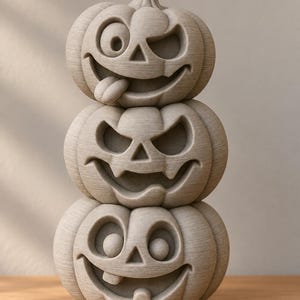May include: A stack of three light grey, textured pumpkin sculptures. The top pumpkin has a winking face with a tongue sticking out. The middle pumpkin features a classic carved jack-o'-lantern expression. The bottom pumpkin has a wide, toothy grin.