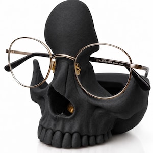 May include: A matte black skull-shaped glasses holder featuring a textured surface. The skull is designed to hold a pair of round-framed glasses with thin gold-toned metal rims and black temple tips. The text "VIOLISTS HSK" is printed on the frame.