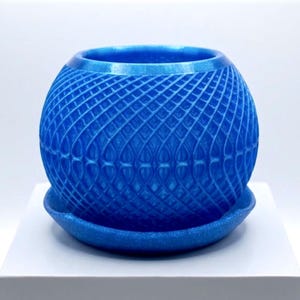 Puede incluir: Una maceta esférica azul con un acabado brillante y con purpurina, acompañada de un plato circular a juego. El exterior de la maceta presenta un patrón de celosía intrincado en relieve que crea un diseño geométrico texturizado.
