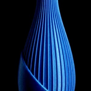 May include: A vibrant blue vase featuring a textured, ribbed surface with vertical lines running along its elongated, tapered body. The design includes a smooth, curved outer layer that partially overlaps the main ribbed structure, creating a layered effect.