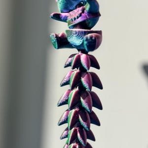 Op de afbeelding: Een 3D-geprint, beweeglijk berenfiguur in iriserende kleuren blauw, paars en groen. De beer zit, met een gesegmenteerde, bloemachtige staart die naar beneden uitsteekt. Het speelgoed hangt tegen een neutrale achtergrond.