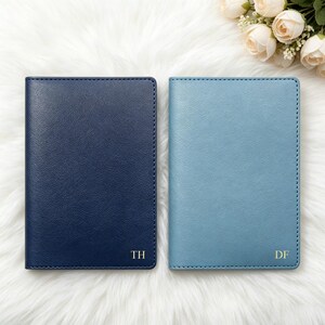 Puede incluir: Se muestran dos fundas de pasaporte de cuero, una azul marino y otra azul claro. Cada una tiene un borde cosido e iniciales doradas. La funda azul marino tiene "TH" y la azul claro tiene "DF". El fondo es una superficie blanca y esponjosa.