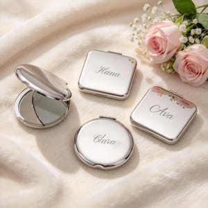 May include: Three polished silver-tone metal compact mirrors with hinged lids. Two are square with the names Hana and Ava engraved, and one is round with the name Clara engraved. One additional round mirror is shown open to reveal the reflective surface.