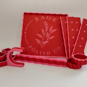 May include: A collection of red and pink accessories including a square tray with the text SAGE POLISH CO. and a floral graphic, three small square dishes, a C-shaped stand, a long wavy strip, three rectangular panels with dots, and two round bowls.